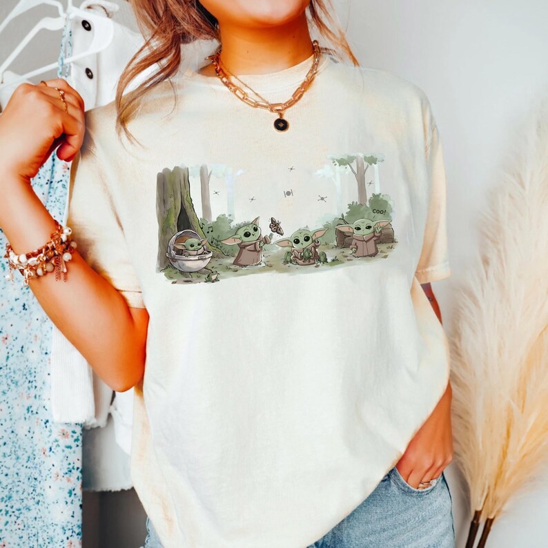 Puede incluir: Camiseta color crema con una escena de bosque con personajes de Grogu. El gr&aacute;fico incluye a Grogu en una c&aacute;psula, con una mariposa y cerca de un &aacute;rbol. La palabra "cool" est&aacute; impresa en un tronco. La camiseta la lleva una persona.