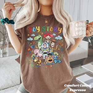 May include: Espresso-coloured t-shirt featuring a colourful graphic of Toy Story characters. The words "TOY STORY" are written across the top in rainbow colours. The shirt is a Comfort Colors brand.