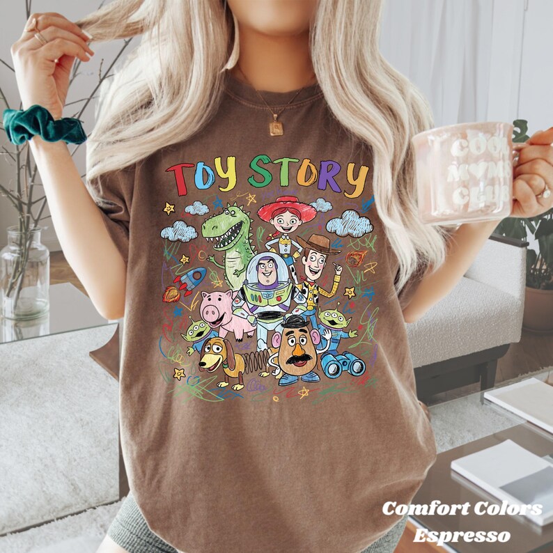 May include: Espresso-coloured t-shirt featuring a colourful graphic of Toy Story characters. The words "TOY STORY" are written across the top in rainbow colours. The shirt is a Comfort Colors brand.