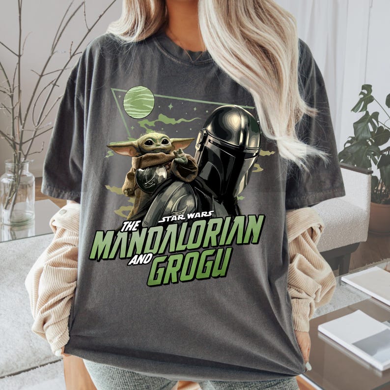 May include: A charcoal gray t-shirt featuring a graphic print of The Mandalorian and Grogu. The design includes a green planet, a triangular background, and the text "STAR WARS THE MANDALORIAN AND GROGU" in bold green and white lettering.