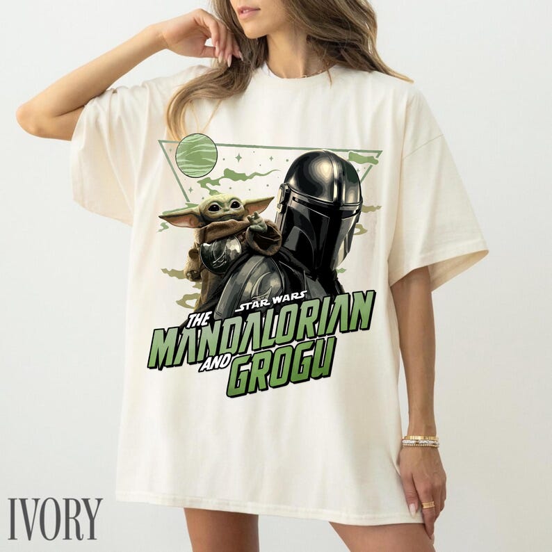May include: An ivory short-sleeve t-shirt featuring a graphic print of The Mandalorian and Grogu. The design includes a green planet, a triangular frame, and the text "STAR WARS THE MANDALORIAN AND GROGU" in bold green lettering.