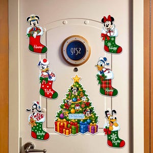 May include: A set of six Christmas stockings featuring Disney characters Mickey, Minnie, Daisy, Donald, Goofy, and Pluto, each with a name space, alongside a central Christmas tree graphic with presents and the text The Smith Family.
