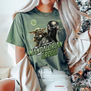 May include: A sage green short-sleeve t-shirt featuring a graphic print of The Mandalorian and Grogu. The design includes the text "STAR WARS THE MANDALORIAN AND GROGU" in green lettering, a small planet, and a triangular background element.