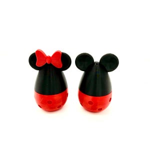 May include: Two novelty salt and pepper shakers. One shaker features a red bow, black ears, and a red base with circular cutouts. The other shaker has black ears and a red base with circular cutouts. Both shakers have a black upper body.