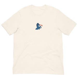 May include: Cream-colored t-shirt featuring a blue horse with red laser beams shooting from its eyes. The horse is in a rearing position. The shirt has short sleeves and a crew neck.