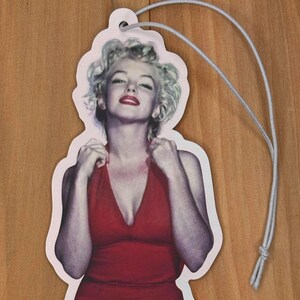 May include: A white car air freshener featuring a photograph of a woman in a red dress. The woman has blonde hair and is looking at the camera.