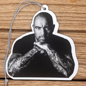 Puede incluir: Ambientador en blanco y negro con un retrato de un hombre con tatuajes en los brazos. La imagen está recortada con una forma única y tiene una cuerda gris para colgar. El fondo es una superficie de madera.