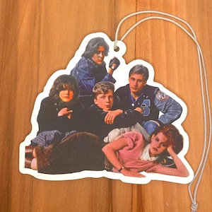 Puede incluir: Un ambientador para coche con un grupo de cinco personas, tres hombres y dos mujeres, de la película de los años 80 The Breakfast Club. Los personajes están vestidos con sus atuendos icónicos de la película. El ambientador tiene forma de corazón y un borde blanco.