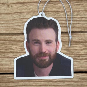 Op de afbeelding: Witte, gestanste luchtverfrisser met een kleurenportret van een man met bruin haar en een baard. De luchtverfrisser heeft een touwtje om op te hangen. De achtergrond is een houten oppervlak.