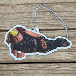 May include: A novelty air freshener featuring a celebrity in a black outfit with a studded belt and wristbands, and a yellow and black hairstyle. The air freshener has a white string for hanging.