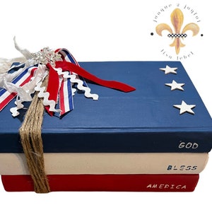 May include: A stack of three decorative books painted in red, white, and blue, with the words "GOD BLESS AMERICA" on the spines. The top book is navy blue with white stars. A decorative bow with red, white, and blue ribbons and a silver embellishment is tied around the books with twine.