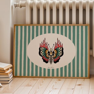 May include: A framed print with a butterfly design. The butterfly is set in an oval shape with a cream background, against a green and white striped background. The butterfly has red, black, and green details. The frame is light brown.