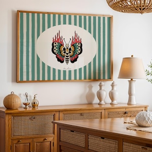 May include: A framed artwork featuring a butterfly design with flames, set against a background of vertical teal and white stripes. The butterfly has black, red, and teal details. The artwork is displayed above a wooden cabinet.