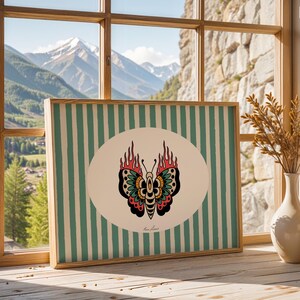 May include: Framed artwork with a butterfly design and flames, set against a striped background. The artwork is displayed on a wooden surface near a window with a mountain view. Two vases with dried plants are also visible.