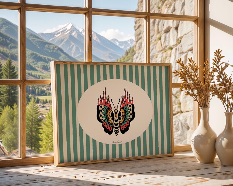 May include: Framed artwork with a butterfly design and flames, set against a striped background. The artwork is displayed on a wooden surface near a window with a mountain view. Two vases with dried plants are also visible.
