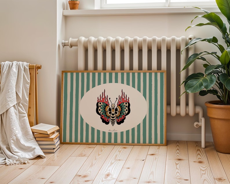 May include: A framed print with a butterfly design. The butterfly is set in an oval shape with a cream background, against a green and white striped background. The butterfly has red, black, and green details. The frame is light brown.