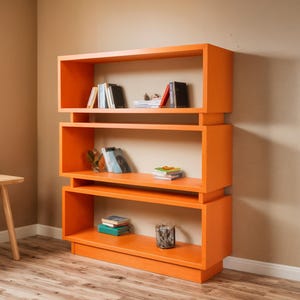 Puede incluir: Una estantería naranja moderna con tres compartimentos abiertos. Las estanterías contienen libros y objetos decorativos. La estantería es de madera y tiene un diseño geométrico. La habitación tiene una paleta de colores neutros con suelo de madera.