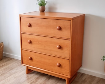 May include: A three-drawer wooden dresser with a light brown finish. The dresser has round wooden knobs and a simple, classic design. A small potted plant sits on top. The dresser is approximately 36 inches wide, 30 inches tall, and 18 inches deep.