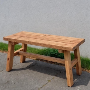 May include: A natural wood bench with a rectangular top and four sturdy legs. The bench is made of light brown wood and has a simple, rustic design. The bench is approximately 48 inches long, 16 inches wide, and 18 inches high.