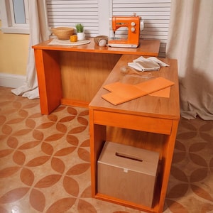 Puede incluir: Una máquina de coser naranja está sobre una mesa de madera. Al lado hay una mesa más pequeña, con una caja de madera debajo. Las mesas están en una habitación con suelo estampado y una ventana.
