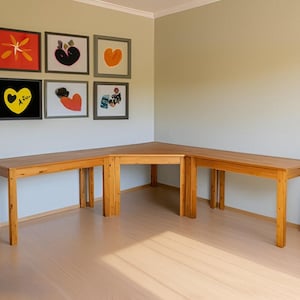 Könnte beinhalten: Ein Ecktisch aus hellem Holz mit drei Teilen. Der Tisch steht in einem Raum mit einer hellbeigen Wand und sechs gerahmten Bildern mit bunter abstrakter Kunst.