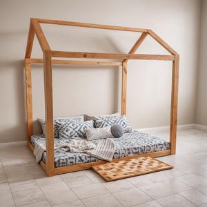 Op de afbeelding: Een houten bedframe in de vorm van een huis met een matras en kussens. Het bed heeft een matrasovertrek met patroon, decoratieve kussens en een klein houten kleed. Het frame is gemaakt van lichtgekleurd hout.