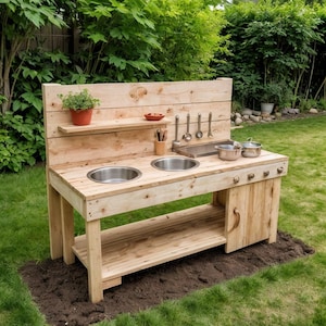May include: A wooden outdoor play kitchen with two stainless steel sinks, a shelf, and a backsplash. It includes pots, utensils, and a small plant. The play kitchen is set on a patch of dirt in a grassy backyard.