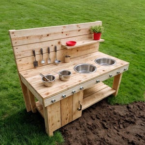 May include: A wooden outdoor play kitchen with a natural finish. It features two stainless steel sinks, a shelf, and hanging utensils. A small red bowl and a potted plant add color. The kitchen is set on a grassy lawn.