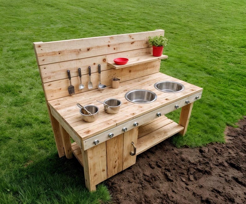 May include: A wooden outdoor play kitchen with a natural finish. It features two stainless steel sinks, a shelf, and hanging utensils. A small red bowl and a potted plant add color. The kitchen is set on a grassy lawn.