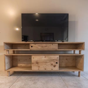 Puede incluir: Un mueble de TV de madera clara con un televisor negro grande encima. El mueble tiene estanterías abiertas y dos cajones. El mueble está sostenido por patas cortas y redondeadas. La veta de la madera es visible.