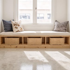 Peut inclure: Banquette en bois sous une fenêtre, avec un coussin crème et trois paniers de rangement en osier. Deux coussins décoratifs à motifs de feuilles et rayures sont posés contre la fenêtre. Le banc est en bois clair.