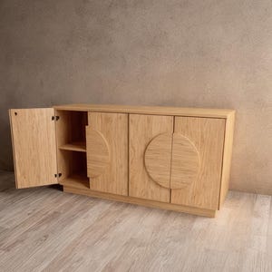 Op de afbeelding: Een licht eikenhouten kast met twee deuren en twee laden. Eén deur is open en onthult een plank. De kastdeuren hebben een halve cirkel ontwerp. De kast staat tegen een neutraal getinte muur en een lichte houten vloer.