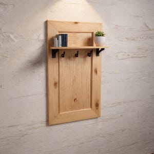 May include: A wooden wall-mounted shelf with a small shelf, three hooks, and a rectangular design. The shelf holds a small potted plant and several books. The wood has a natural finish, and the hooks are black.