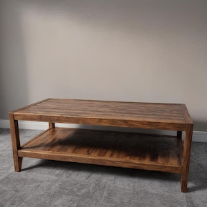 May include: A rectangular, two-tiered coffee table made of dark brown wood. The table has a lower shelf for storage and a simple, modern design. The dimensions are approximately 48 inches long, 24 inches wide, and 18 inches high.