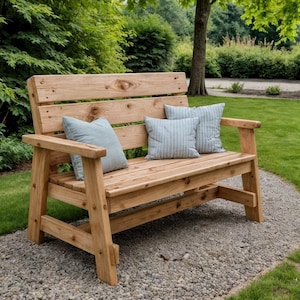 Könnte beinhalten: Eine Gartenbank aus Holz mit natürlicher Oberfläche, mit Lattenrücken und Armlehnen. Zwei hellblaue Kissen mit weißen Streifen liegen auf der Sitzfläche. Die Bank steht auf einem Kiesweg, umgeben von Grün.