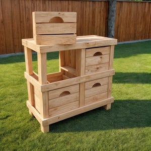 Może przedstawiać: Jasna drewniana jednostka do przechowywania z szufladami i górną półką oraz małą drewnianą skrzynią na górze. Jednostka ma naturalne wykończenie słojów drewna i stoi na zielonym trawniku.