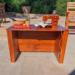 May include: A wooden sewing table with a vintage orange sewing machine. A wooden bowl, orange, red cloth, and potted plant are on the table. The table is made of reddish-brown wood and has a simple design.
