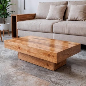 May include: A rectangular wooden coffee table with a light brown finish. The table has a solid, blocky design with a lower base. The table is in front of a beige sofa with matching pillows. The floor is tiled.
