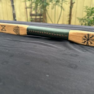 May include: A wooden bow with a dark green leather grip and etched symbols. The bow has a natural wood grain finish and is designed for archery. The grip is stitched with brown thread.