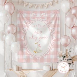 May include: A pink and white checkered backdrop with the text "AMELIA IS ONE silly goose" in a decorative frame. The backdrop is decorated with balloons, a banner, and a floral arrangement. Two small gift boxes are on the table.