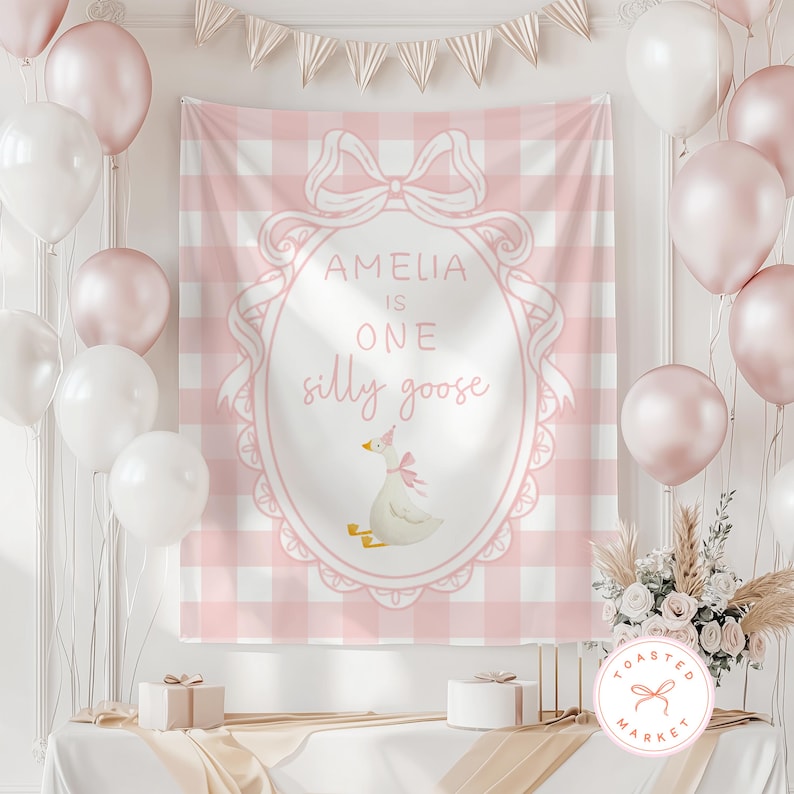 May include: A pink and white checkered backdrop with the text "AMELIA IS ONE silly goose" in a decorative frame. The backdrop is decorated with balloons, a banner, and a floral arrangement. Two small gift boxes are on the table.