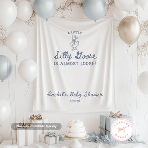 Pu&ograve; includere: Un arazzo bianco con la scritta "A Little Silly Goose Is Almost Loose!" &egrave; il punto focale di questa immagine. L'arazzo &egrave; decorato con un'oca dei cartoni animati e le parole "Rachel's Baby Shower" e la data 5.29.26. Palloncini bianchi e blu circondano l'arazzo.