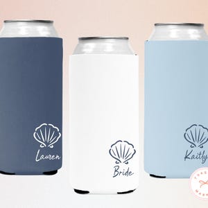 May include: Three can coolers in navy, white, and light blue. Each cooler has a white seashell graphic and a name or title: "Lauren", "Bride", and "Kaitlyn". The coolers are designed to keep beverages cold.