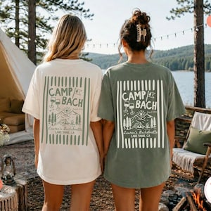 May include: Two short-sleeve t-shirts, one ivory and one moss green, each featuring a rectangular graphic on the back with vertical stripes, pine trees, a tent, a sun, wine glasses, and the text CAMP BACH, Lauren's Bachelorette, LAKE TAHOE, CA.