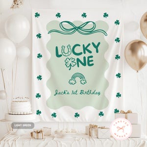 Puede incluir: Una pancarta blanca con un diseño ondulado verde claro, tréboles verdes y el texto "LUCKY ONE" con una herradura, un trébol y un arcoíris. También está presente el texto "Jack's 1st Birthday". La pancarta es parte de una fiesta de cumpleaños.