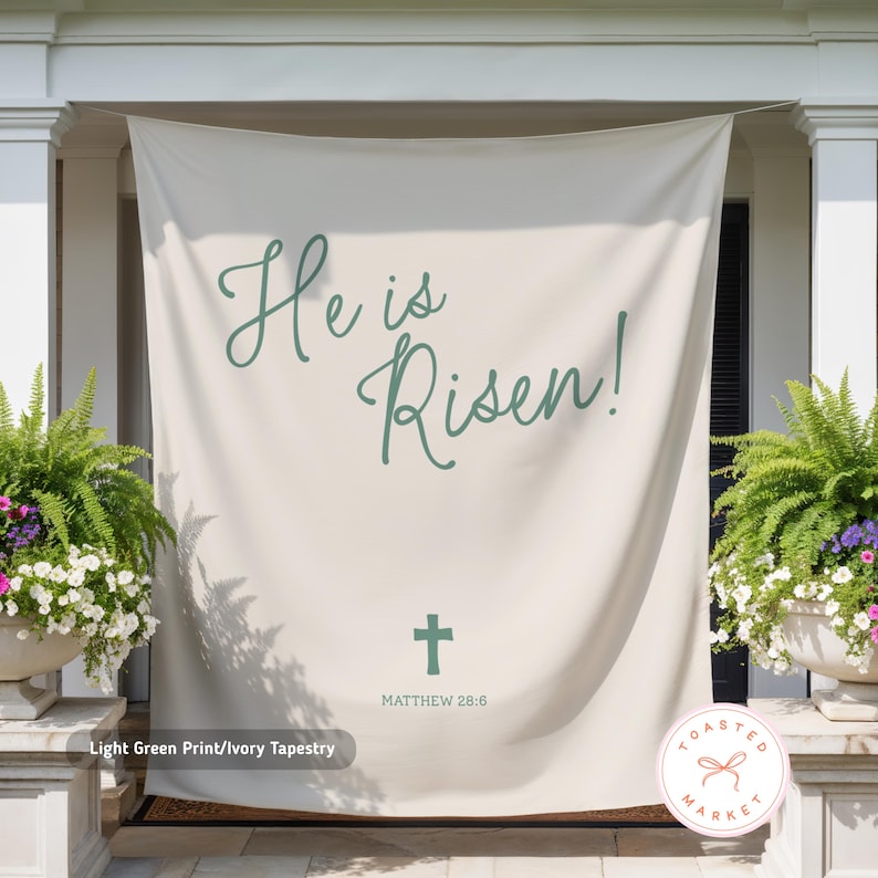 May include: Ivory tapestry with the text "He is Risen!" in light green script, a green cross, and "Matthew 28:6." The tapestry is displayed outdoors, flanked by potted plants.