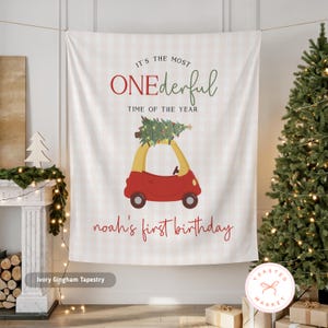 May include: Ivory gingham tapestry with the text "IT'S THE MOST ONEderful TIME OF THE YEAR" and "noah's first birthday". A red and yellow car with a Christmas tree on top is in the center. The tapestry is hanging in front of a Christmas tree.