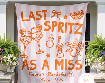 May include: A large rectangular fabric banner with vertical light pink and white stripes. The banner features orange graphic illustrations of two cocktail glasses, a diamond ring, a bottle, and citrus slices, with the text LAST SPRITZ AS A MISS, Emma's Bachelorette, and Miami 2026.