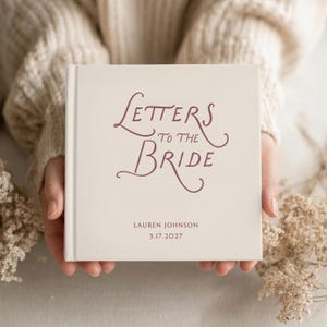 Puede incluir: Un libro de tapa dura de color crema con el texto "Letters to the Bride" en una caligrafía cursiva de color granate oscuro. Debajo del título, el nombre "Lauren Johnson" y la fecha "17.03.2027" están impresos en una fuente sans-serif sencilla.