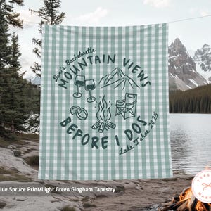 May include: A light green gingham tapestry with the text "Mountain Views Before I Dos" and illustrations of wine glasses, rings, a campfire, and chairs. The tapestry is hanging outdoors with a lake and mountains in the background.